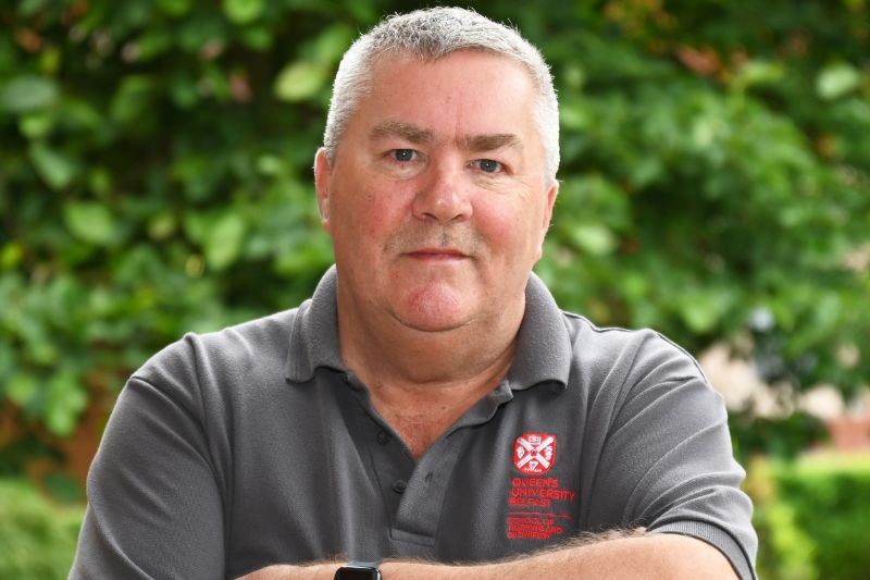 Grey haired man standing crossed armed wearing a short sleeved grey Queen's university polo shirt, standing in front of some shrubbery.